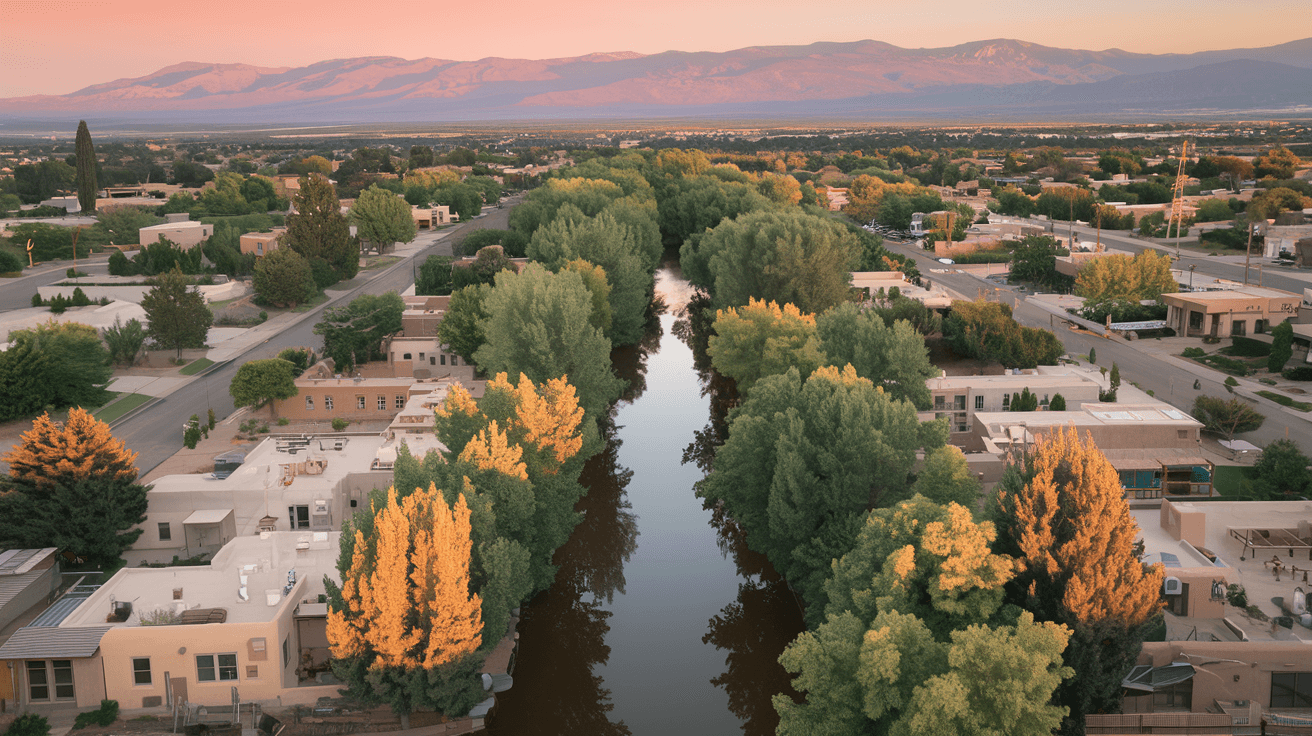 Aerial view of an Albuquerque neighborhood at golden hour, with adobe-style homes, mature cottonwood trees along an acequia, and the Sandia Mountains glowing pink in the background