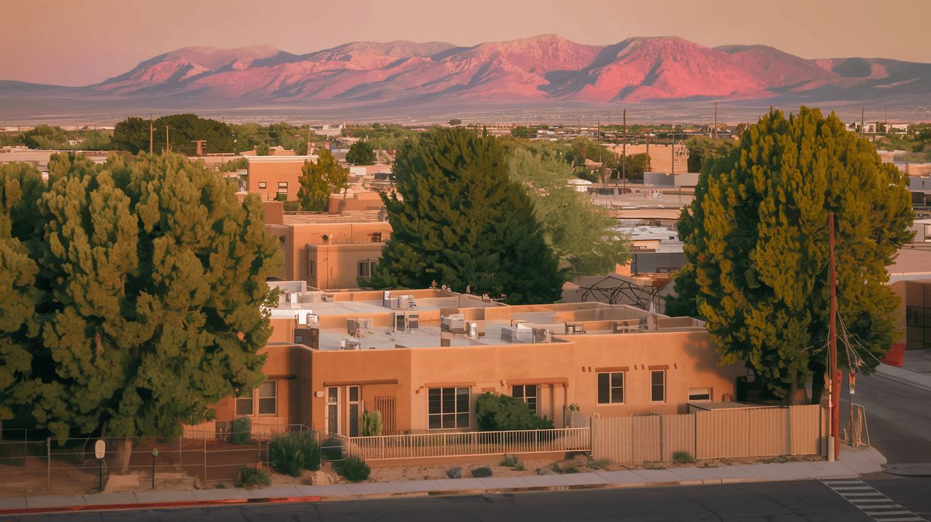 Aerial view of an Albuquerque residential neighborhood at golden hour with the Sandia Mountains glowing pink in the background, adobe rooftops and mature cottonwood trees visible below