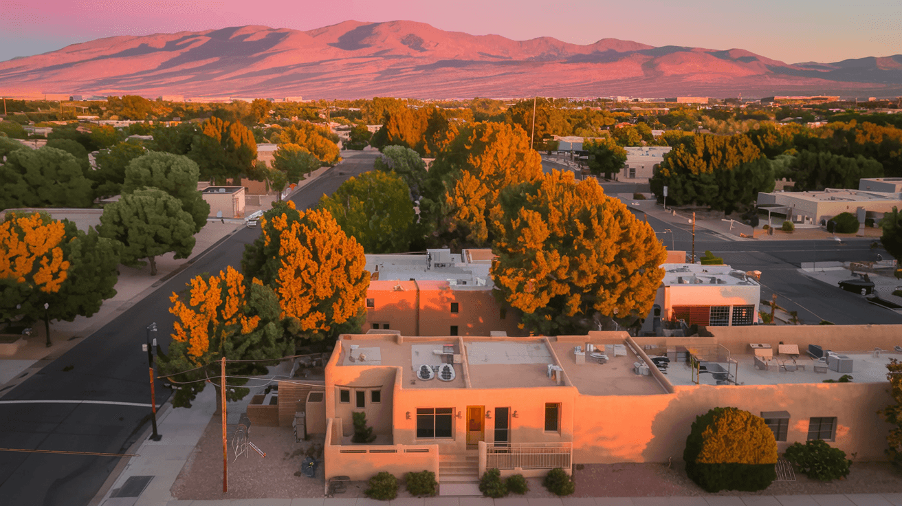 Aerial view of an Albuquerque neighborhood at golden hour with the Sandia Mountains glowing pink in the background, showing tree-lined streets and adobe-style homes